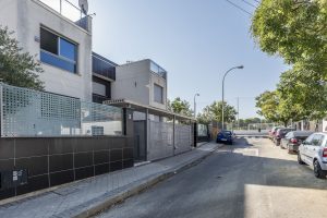 Puerta de entrada de un edificio moderno en un barrio residencial, rodeado de árboles y coches estacionados en la calle.